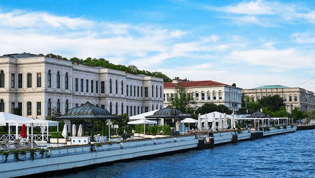 유서 깊은 흰색 건물, 울창한 녹지, 야외 좌석 공간, 구름이 흩날리는 푸른 하늘이 있는 수변 전망.