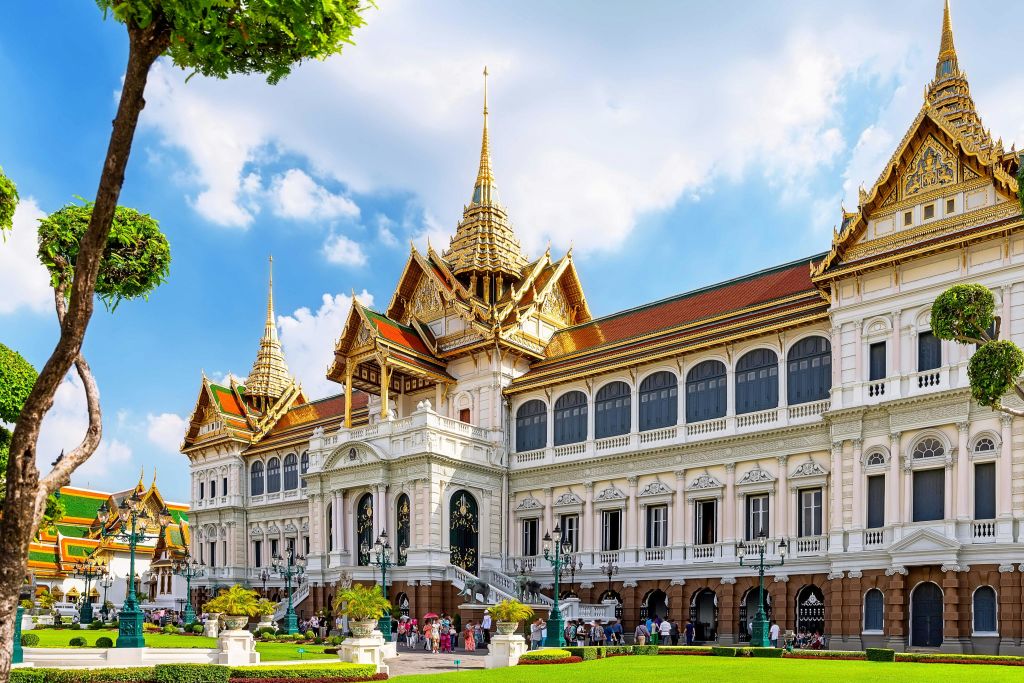 푸른 하늘 아래 관광객과 울창한 녹지로 둘러싸인 황금빛 첨탑과 섬세한 건축물이 있는 화려한 역사적 건물입니다.