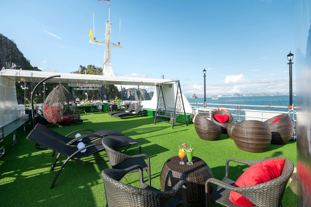 고리버들 가구가 비치된 배의 야외 라운지 공간, 테이블 위의 음료, 행잉 체어, 해안 경치를 감상할 수 있는 맑고 푸른 하늘.