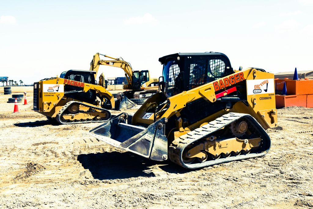 다른 장비를 배경으로 건설 현장에서 "Badger" 와 "Pacman" 이라고 표시된 두 개의 노란색 스키드 스티어 로더.