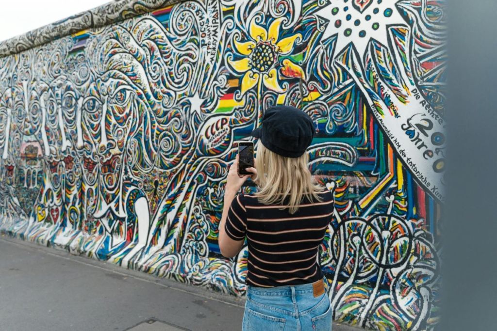 줄무늬 셔츠와 검은 모자를 쓴 여성이 야외 벽에 걸린 화려하고 복잡한 벽화 사진을 찍고 있습니다.