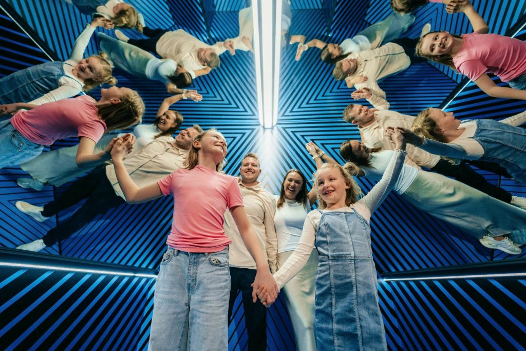 파란색 줄무늬 벽과 밝은 조명이 있는 거울이 있는 방에서 미소를 짓고 손을 잡고 있는 4인 가족.