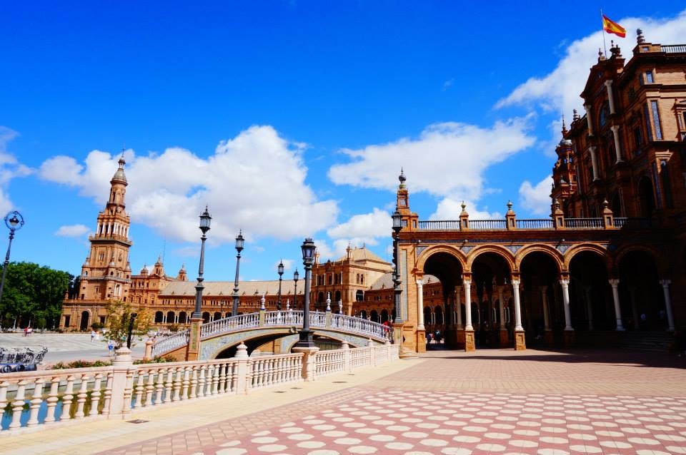 구름이 흩어져 있는 밝고 푸른 하늘 아래, 아치형 건축물, 화려한 가로등 기둥, 다리가 있는 광장.