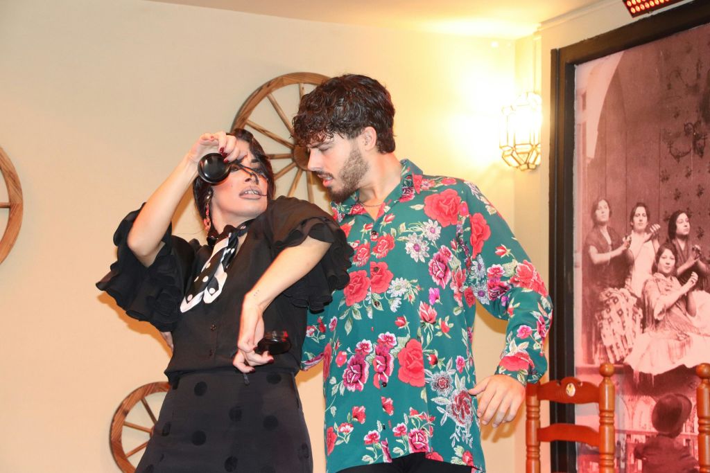 남자와 여자는 열정적으로 춤을 추고, 남자는 꽃무늬 셔츠를 입습니다. 배경에는 액자 사진과 나무 바퀴가 있습니다.