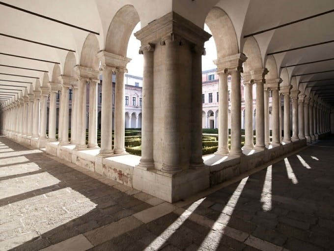 석조 타일 바닥에 그림자를 드리우며 기둥으로 된 아치형 마당입니다. 잘 다듬어진 수풀과 뒷편에 있는 건물.