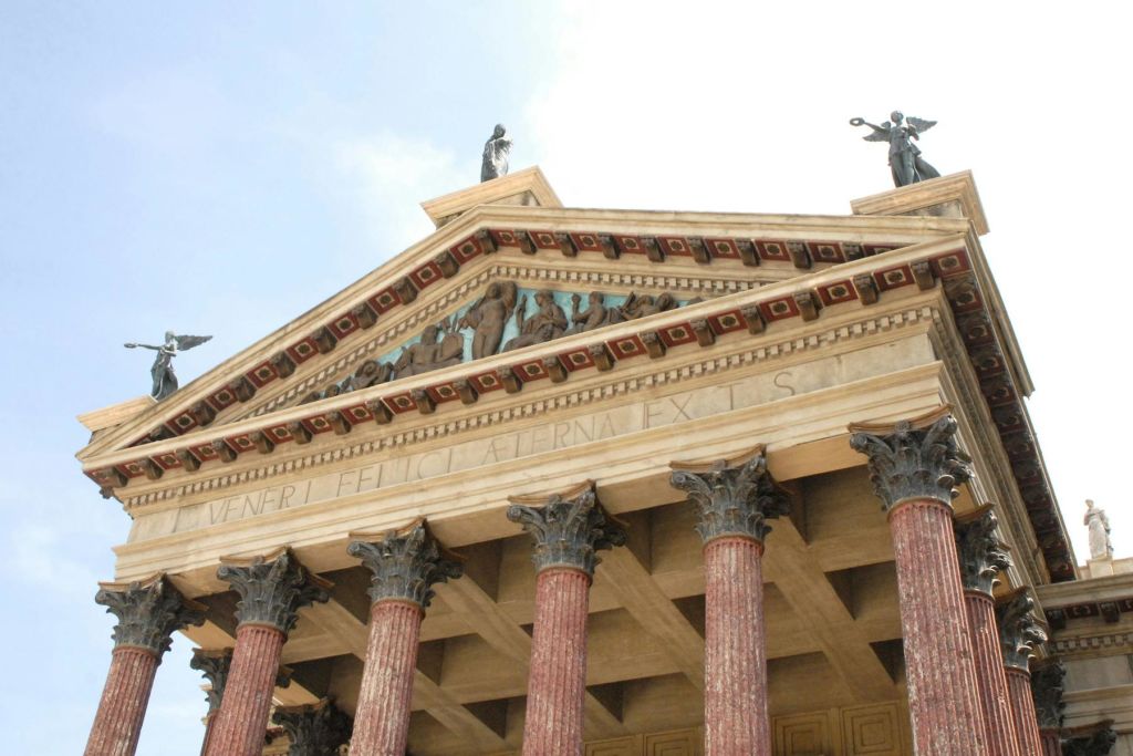 동상, 장식 요소, 세밀한 조각이 있는 페디먼트를 받치고 있는 기둥이 있는 신고전주의 양식의 건물 외관입니다.