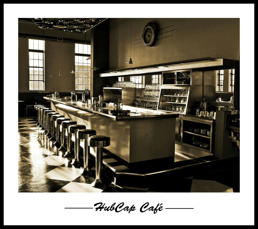 긴 카운터, 크롬 바 스툴, 체크무늬 바닥, 커다란 창문이 있는 복고풍 카페입니다. "HubCap Café" 는 아래에 캡션되어 있습니다.