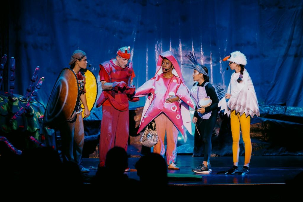 다채로운 색상의 바다 생물 의상을 입은 네 명의 배우가 바다를 테마로 한 소품과 파란색 배경으로 무대에서 공연합니다.