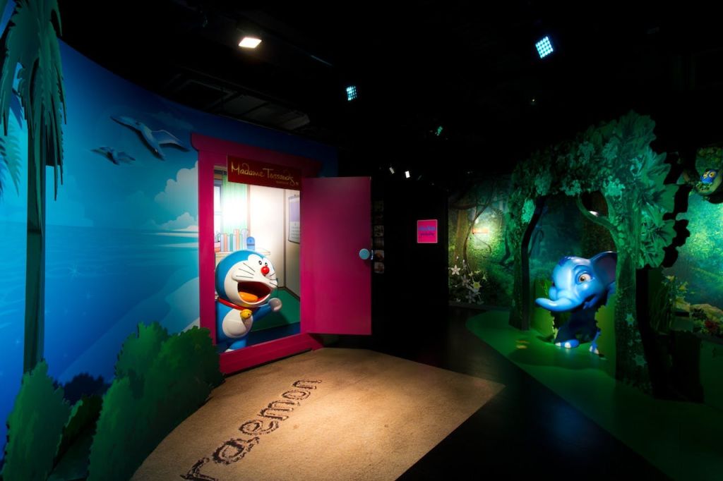 분홍색 문에서 나오는 도라에몽 피규어와 근처에 움직이는 파란색 코끼리가 있는 실내 장면. 다채로운 벽화가 있는 희미한 조명.