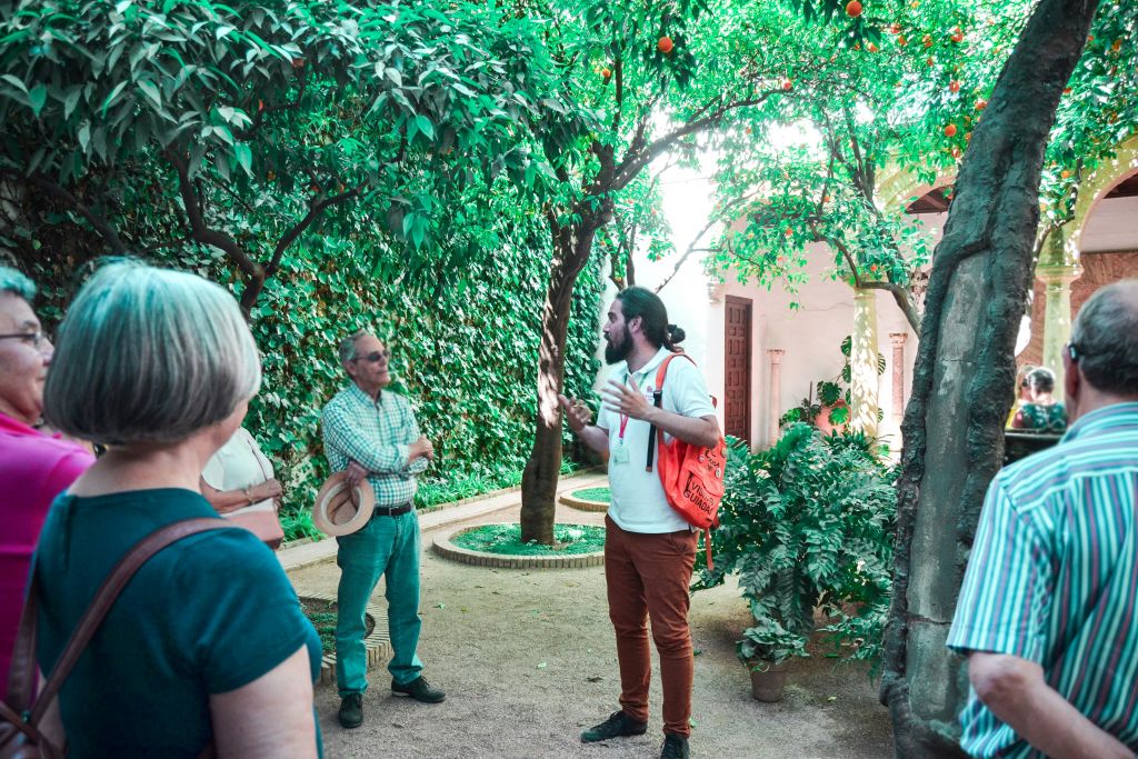 나무와 담쟁이 나무로 덮인 벽이 있는 울창한 정원에서 주황색 배낭을 든 가이드가 소수의 사람들에게 이야기합니다.