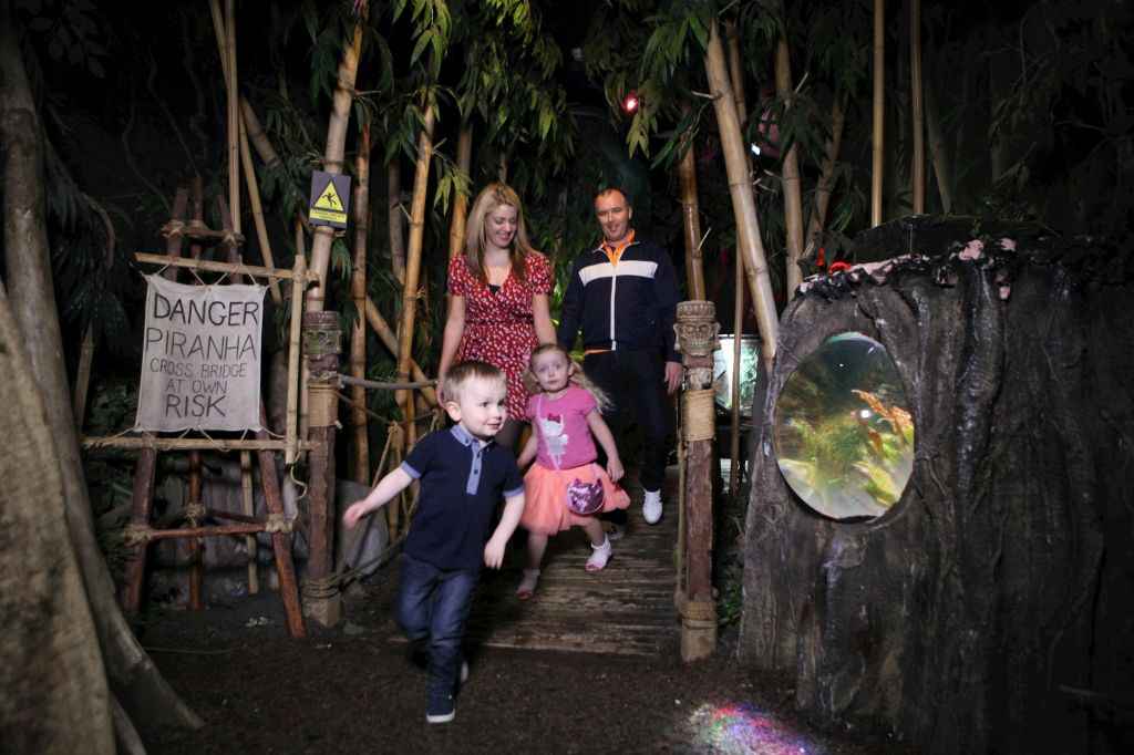 피라냐에 대한 경고 표지판이 있는 정글 테마의 나무 다리에서 어른 두 명을 앞질러 달리는 두 명의 어린이.