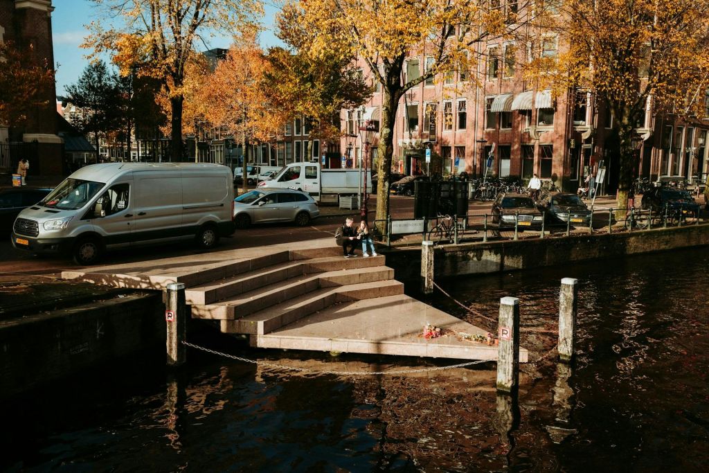 운하 옆 계단에 주차된 자동차, 자전거, 가을 나무를 배경으로 두 사람이 앉아 있습니다.