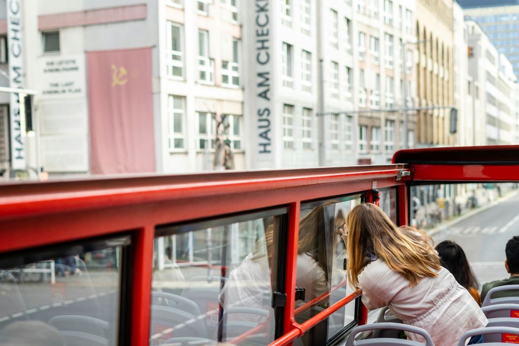 한 여성이 저명한 "HAUS AM CHECKPOINT CHARLIE" 표지판이 있는 도시 지역의 오픈탑 빨간색 버스에서 사진을 찍고 있습니다.