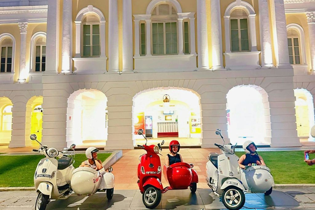 세 사람이 사이드카를 타고 빈티지 스쿠터에 앉아 야간에 조명이 켜진 클래식 스타일의 건물 앞에 주차합니다.