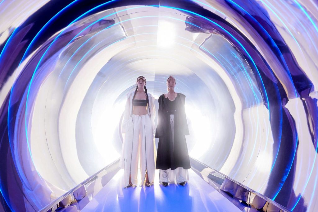 파란색과 흰색 불빛이 반짝이는 터널에 미래형 옷을 입은 두 사람이 서 있습니다.