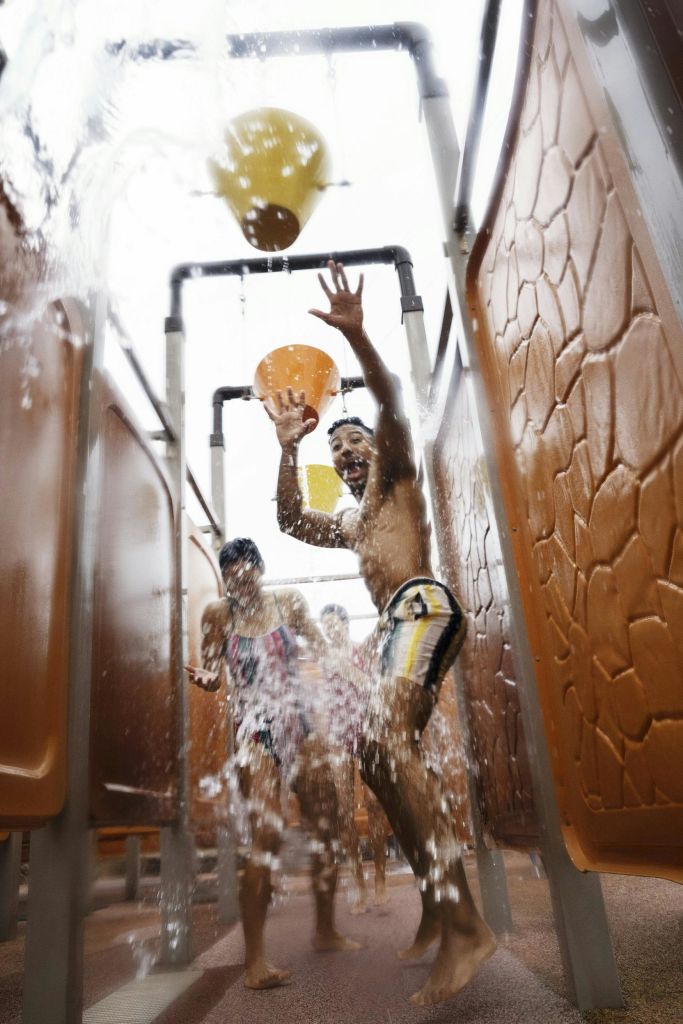 워터 슬라이드의 사람들은 머리 위 양동이에서 물이 튀기려고 하며, 측면과 배경에는 주황색 패널이 있습니다.