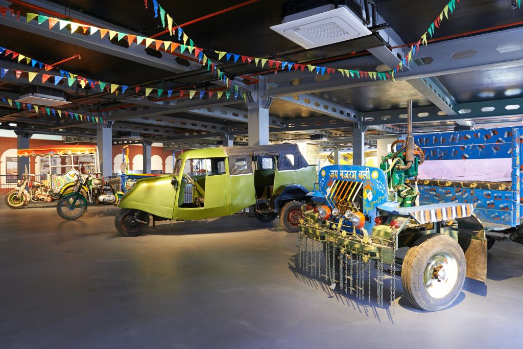 박물관이나 전시 공간의 줄 조명과 깃발 아래 실내에 화려한 오토 릭쇼와 장식된 트랙터가 전시되어 있습니다.