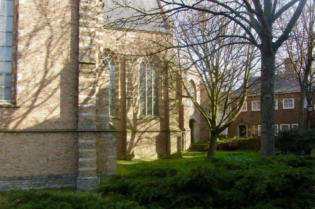 맨발의 나무와 푸른 수풀로 둘러싸인 아치형 창문이 있는 햇살 가득한 벽돌 교회는 잔디와 구조물에 그림자를 드리우고 있습니다.