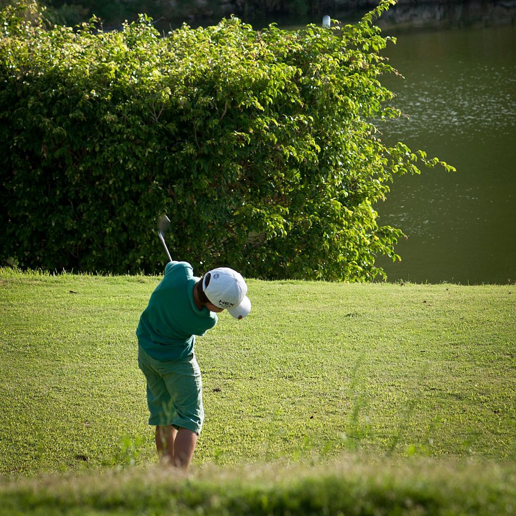 골프를 치고, 호수와 수풀을 배경으로 잔디가 우거진 코스에서 클럽을 휘두르는 초록색 의상을 입은 사람.