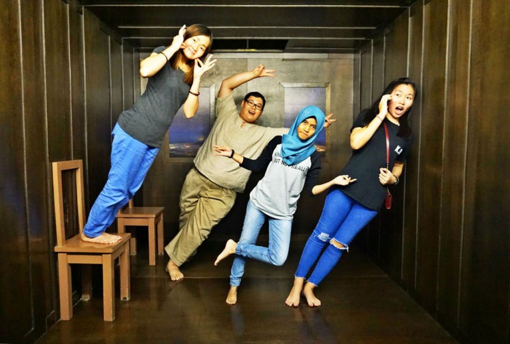 기울어진 방에 네 사람이 서 있습니다. 두 사람은 장난스럽게 포즈를 취하고, 한 명은 미소를 짓고, 한 명은 전화 통화 중 놀란 표정을 짓습니다.