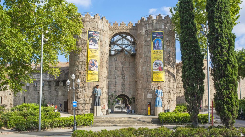 두 개의 탑, 화려한 배너, 동상, 나무, 걸어다니는 사람들이 있는 돌 입구. 배경은 맑은 하늘.