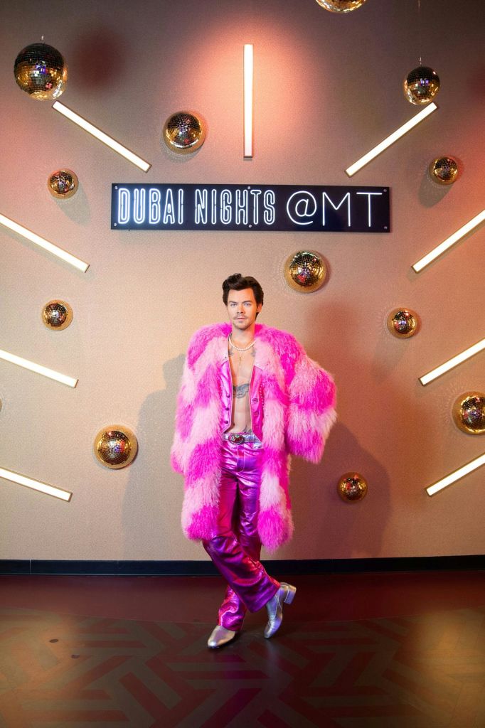 밝은 분홍색 모피 코트와 빛나는 바지를 입은 사람이 조명과 거울로 둘러싸인 "DUBAI NIGHTS @ MT" 라고 적힌 표지판 앞에 서 있습니다.