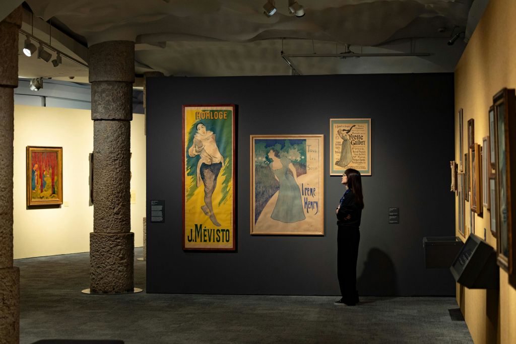 한 사람이 아트 갤러리의 검은 벽에 표시된 빈티지 포스터를 관찰하고 있습니다. 객실은 어두운 조명과 기둥을 갖추고 있습니다.