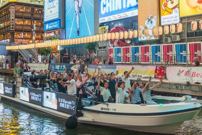 화려한 광고판이 있는 분주한 도시 지역의 조명등 아래로 환호하는 사람들로 가득한 보트가 지나갑니다.