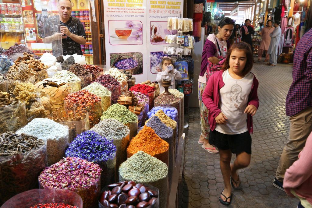실내 시장 가판대에는 다양한 다채로운 향신료와 말린 상품이 전시되어 있으며, 사진 촬영을 하는 남성과 어린이를 포함한 고객들이 둘러보고 있습니다.