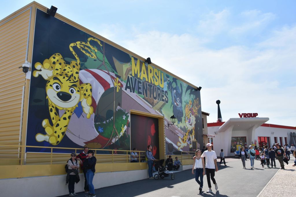 화창한 날에 커다란 "Marsu Aventures" 벽화와 "Vroupe" 입구 표지판이 있는 건물 근처를 걷는 사람들.