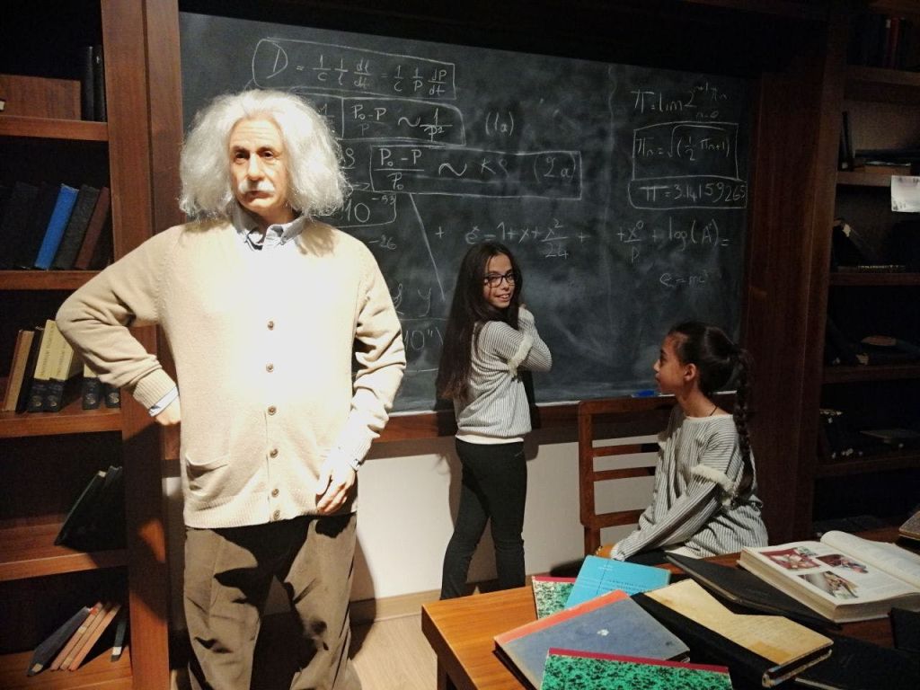 교실 환경에서 알버트 아인슈타인 (Albert Einstein) 을 닮은 왁스 그림 옆에 등식으로 덮인 칠판 옆에 두 아이가 서 있습니다.