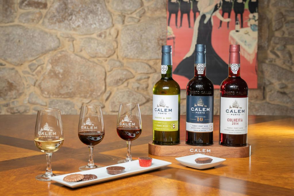 칼렘 포르투 와인 3병, 와인이 가득 담긴 잔, 나무 테이블 위에 배치된 초콜릿이 담긴 접시.