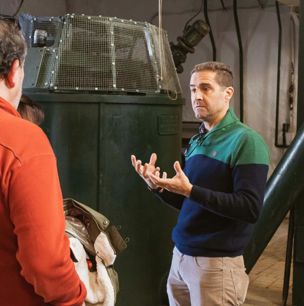 초록색과 파란색 스웨터를 입은 남성이 작업장 환경에서 산업 장비 근처의 두 사람에게 말을 걸고 있습니다.