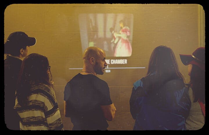 세 사람이 분홍색 드레스를 입은 사람의 이미지가 표시된 화면을 마주보고 '방' 이라는 텍스트와 함께 서 있습니다.