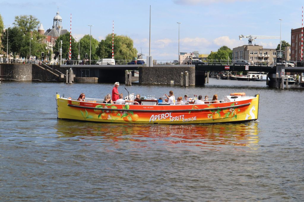 "Aperol Spritz Cruise" 브랜드가 새겨진 노란색 보트는 강을 배경으로 다리와 건물이 있습니다.