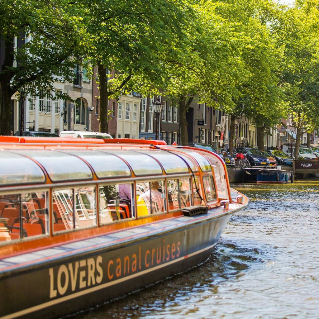 "연인 운하 크루즈" 문구가 적힌 운하 보트가 나무가 우거진 운하를 미끄러지듯 지나가며 주차된 자동차와 주택을 배경으로 펼쳐집니다.