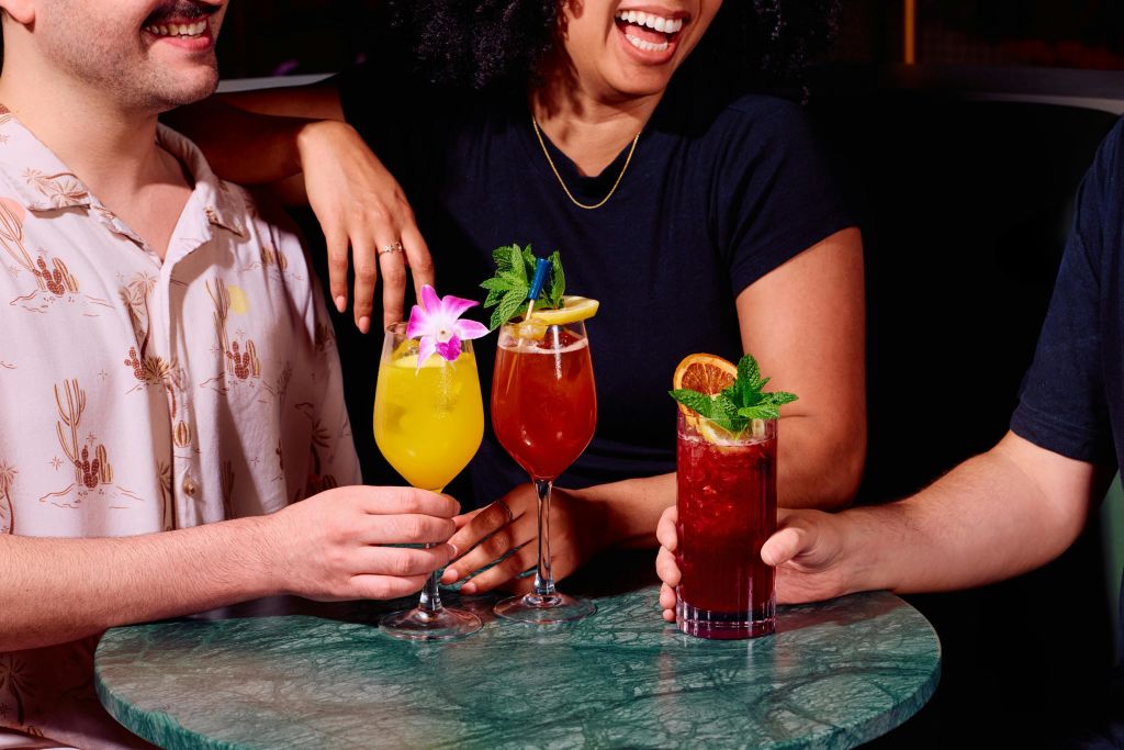 초록색 대리석 테이블에서 꽃, 과일, 민트로 장식된 다채로운 칵테일을 즐기는 세 사람.