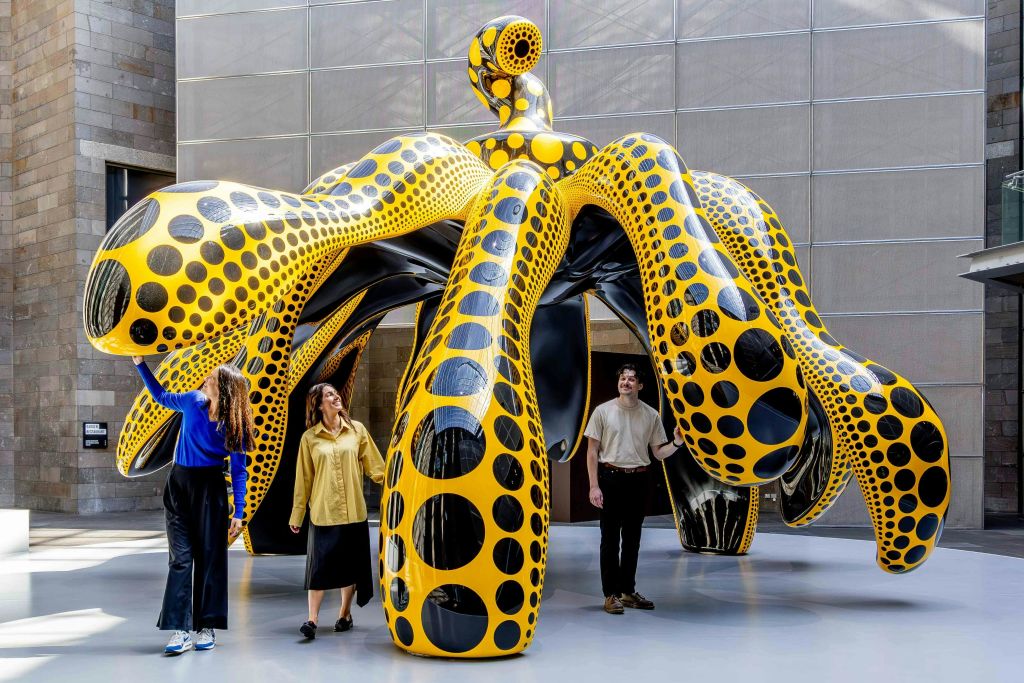 세 사람이 문어를 닮은 커다랗고 화려한 물방울 무늬의 설치물과 상호작용하며, 실내에 전시되어 있습니다.