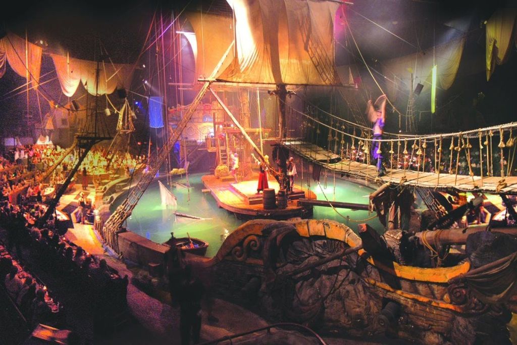 배우들이 의상, 밧줄, 돛, 물, 무대 조명으로 극적인 분위기를 연출하는 연극 해적선 장면.