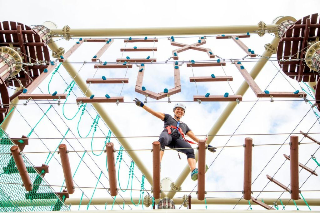 한쪽 팔을 쭉 뻗은 채 미소를 지으며 고가 로프 코스를 항해하는 하네스의 사람. 구조물과 로프가 보입니다.