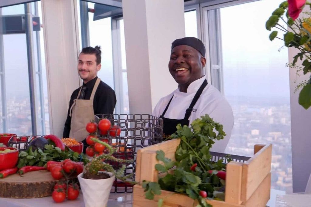 도시가 내려다보이는 커다란 창문을 배경으로 두 셰프가 신선한 채소와 허브가 가득한 주방 카운터에서 미소를 짓고 있습니다.