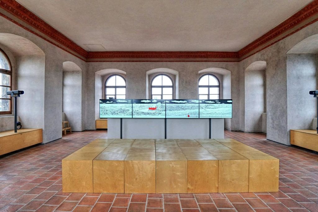 타일 바닥, 3개의 아치형 창문, 벽걸이형 멀티 스크린 디스플레이, 전경에 목재 블록 구조가 있는 객실입니다.