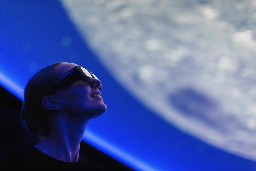 푸른 빛이 비추는 공간의 이미지를 보여주는 대형 스크린을 올려다보는 3D 안경을 쓴 사람.