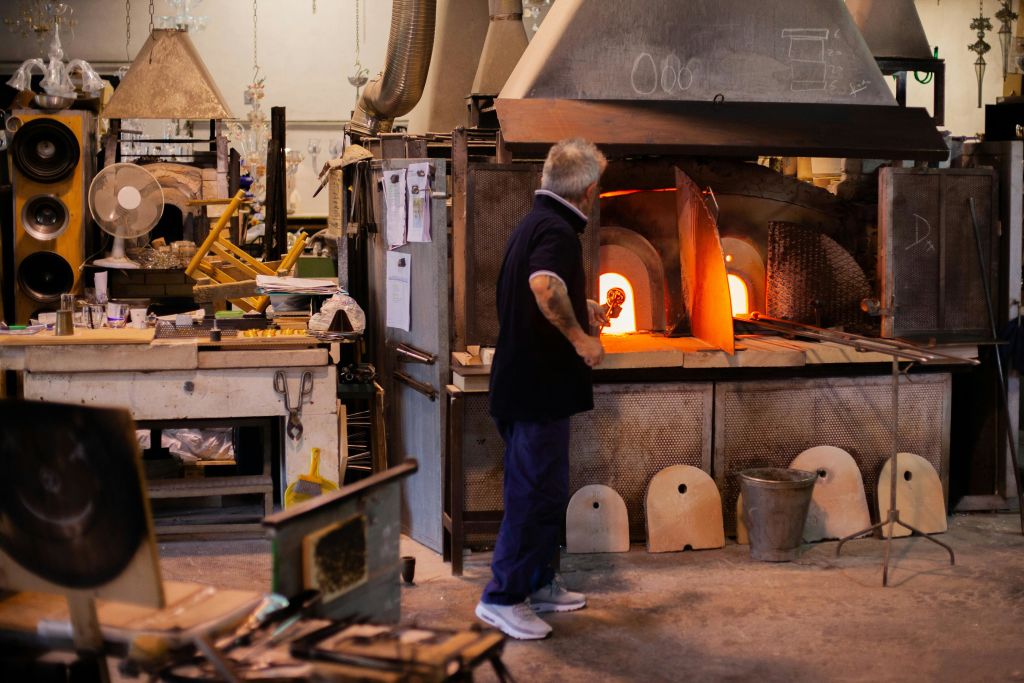 한 남성이 공방의 유리로 앞에 서서 용융 유리가 달린 파이프를 들고 있습니다. 도구와 장비가 그를 둘러싸고 있습니다.