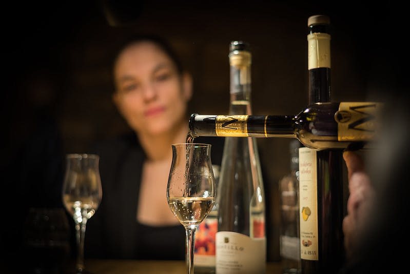 병에 담긴 화이트 와인이 가득 담긴 잔에 초점을 맞춘 배경의 인물. 근처에 다른 잔과 병 두 개가 있습니다.