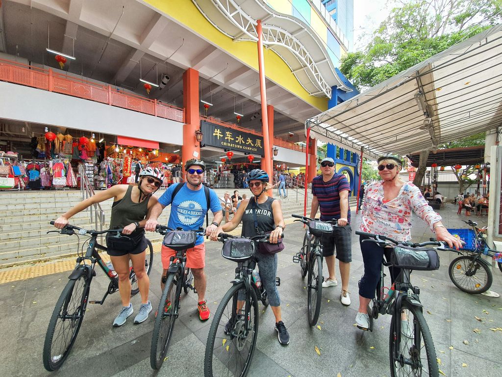 자전거를 탄 네 명이 상점과 간판이 있는 화려한 상업 건물 앞에서 헬멧을 쓰고 미소를 짓고 포즈를 취하고 있습니다.