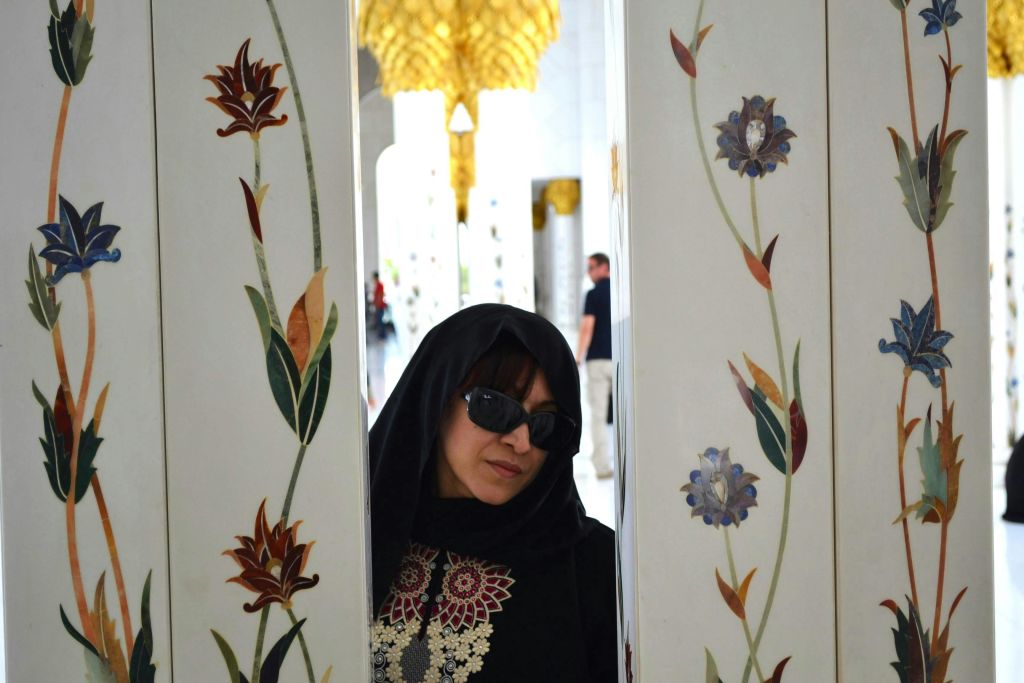 선글라스와 스카프를 두른 여성이 꽃무늬가 있는 화려하게 장식된 기둥 사이에 서 있습니다.