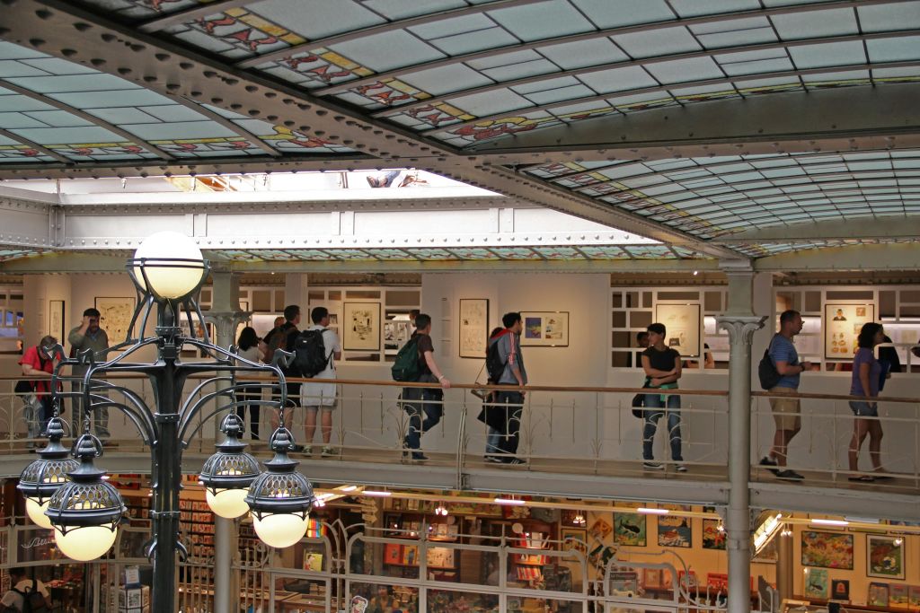 밝은 실내 공간의 발코니에 서 있는 사람들로, 벽에 스테인드글라스 천장과 액자에 담긴 예술품이 있습니다.