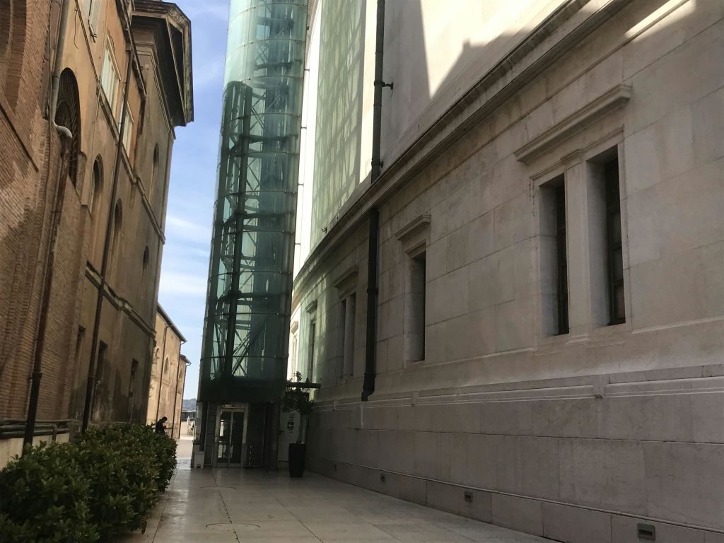 왼쪽에는 유리 엘리베이터 샤프트, 오른쪽에는 창문, 멀리에는 수풀이 있는 건물 사이의 좁은 골목.