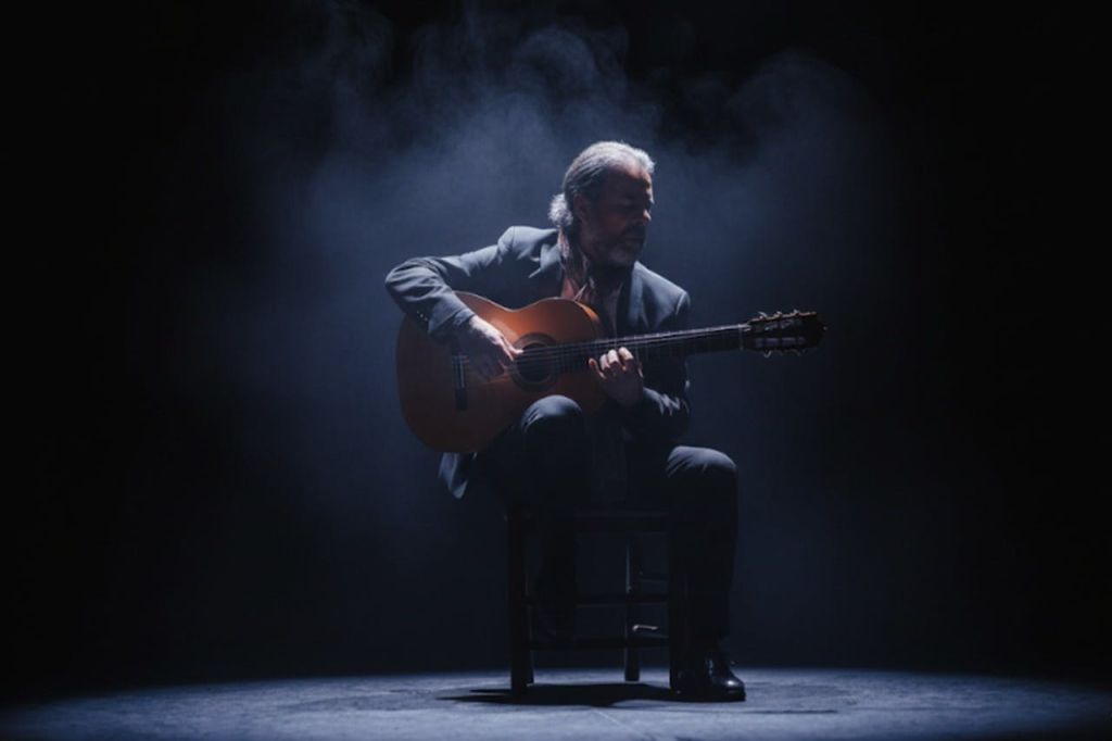 정장을 입은 남성이 의자에 앉아 연기가 자욱한 배경의 드라마틱한 조명 아래에서 어쿠스틱 기타를 연주하고 있습니다.